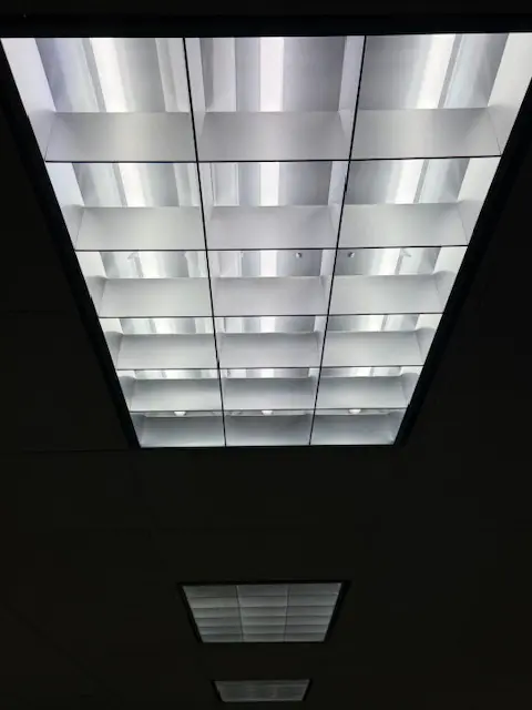 Parabolic louvered fluorescent ceiling light fixture showing high-glare industrial lighting that lacks biological spectral integrity and creates flicker.